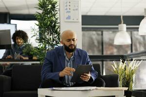 Arab executive manager checking financial data on tablet while working in business office. Startup company employee browsing internet on digital device and sitting on couch in coworking space photo