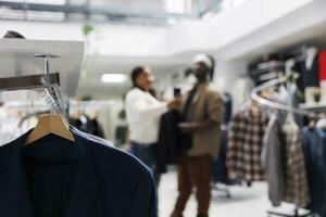 Clothes hanging on rack in store with bloggers taking selfie for brand promotion on blurred background. Casual apparel from new collection on hangers in fashion boutique close up selective focus photo