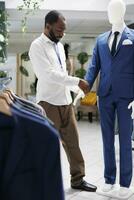 Assistant adjusting jacket on mannequin wearing formal outfit in clothing store. African american man checking male suit on model showcasing apparel for customers in shopping mall photo