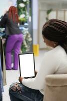 africano americano mujer utilizando tableta burlarse de arriba mientras compras en Moda boutique. cliente participación digital artilugio con blanco vacío pantalla en vertical modo mientras sentado en Departamento Tienda foto