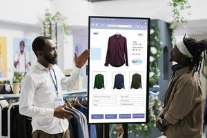 Male assistant showing clothes on interactive kiosk board to young trendy customer in clothing store at mall. Retail employee helping client to choose modern fashion items, self ordering. photo