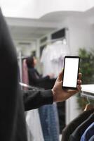 Customer in retail store using mobile phone empty white digital display while checking available apparel options. Shopping mall boutique client showing smartphone blank touchscreen photo