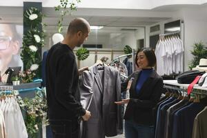 Shopping mall boutique smiling asian woman seller assisting arab man client in selecting jacket. Clothing store worker holding formal apparel hanger, showing outfit to customer, giving fashion advice photo