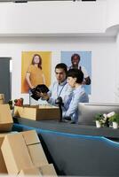 Store workers putting trendy clothes in cardboard boxes, preparing packages for delivery in modern boutique. Diverse team discussing customers orders, checking shipping report on computer photo