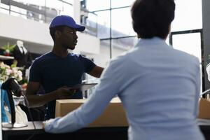 africano americano mensajero explicando Envío detalles a obrero, tomando cardbord cajas desde mostrador escritorio en compras centro comercial. gerente preparando paquetes para entrega, trabajando en moderno boutique foto