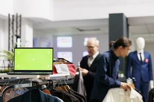 Open laptop computer with chroma key green screen standing on cloth rack in clothing shop, people shopping on background. Small retail business and inventory management software photo