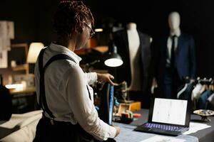 African american couturier measuring sleek bespoken elegant garment for upcoming customer comission. Tailor checking length size of unfinished sartorial business suit in atelier shop workspace photo