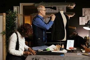 Elderly professional couturier making adjustments on exquisite handmade suit blazer on mannequin while assistant crafts elegant pants. Senior suitmaker and apprentice manufacturing bespoken garments photo