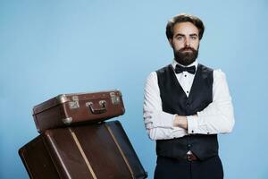 Serious professional bellboy in studio, posing with suit and white gloves over blue background. Hotel concierge standing next to pile of suitcases and luggage to carry, doorman assistance. photo