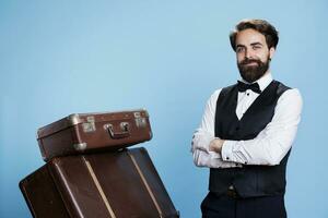 Hotel worker with baggage pile on camera, providing luxury service to guests and helping with suitcases. Professional bellhop or porter dressed in suit and tie feeling determined. photo