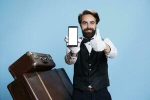 Classy hotel concierge next to luggage holding phone with white empty display, tourism concept. Bellboy showing blank screen on camera, commitment of his professional duties. photo