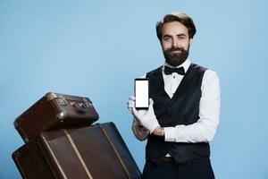 Classy doorman shows white screen on smartphone, holding device while he is in professional hospitality attire. Elegant determined bellboy presents blank copyspace template on display. photo