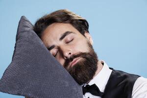 de buen tono botones que cae dormido en almohada mientras él soportes en estudio, sensación extremadamente desgastado fuera y exhausto. profesional empleado portero tomando un rápido siesta en cámara, expresando agotamiento foto