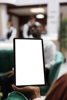 Female person looking at white screen on digital tablet, using blank display with copyspace in lounge area at hotel lobby. Young adult holding device with isolated template. Close up. photo