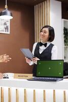 Receptionist having greenscreen on desk, welcoming guests at hotel and starting check in process with tablet. Asian woman using laptop showing isolated display with chroma key copyspace. photo