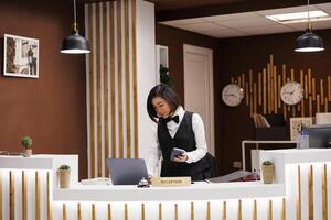 Front desk staff holding pos terminal in hotel lobby, preparing to welcome guests and provide excellent service. Asian receptionist using device for electronic payment at luxury resort. photo