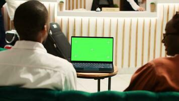 Happy african american tourists watching video on laptop chroma key green screen mock up while sitting in hotel foyer. Guests waiting to be checked in, streaming movie in resort lounge photo