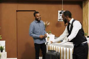 Friendly African American young guy bellhop in uniform helping hotel guest with luggage upon arrival, bellboy smiling and welcoming traveler in lobby assisting in bringing suitcase into room photo
