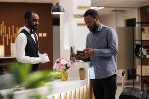 African American guy hotel guest showing his passport to receptionist standing behind reception counter. Traveler with suitcase checking in at front desk upon arrival. People and traveling photo