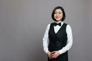 Smiling asian restaurant waitress wearing black and white uniform standing and looking at camera. Cheerful young woman professional receptionist posing for studio portrait photo