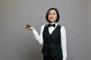 Restaurant receptionist in uniform holding bell in hand, smiling and looking at camera. Catering service cheerful young asian waitress serving and getting attention portrait photo