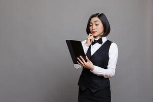 Thoughtful asian waitress in professional uniform managing order on digital tablet. Pensive receptionist thinking while examining restaurant menu on portable gadget and posing in studio photo