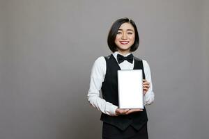 Smiling asian waitress presenting digital tablet empty screen with copy space for advertisement. Friendly restaurant receptionist showing blank touch screen and looking at camera photo