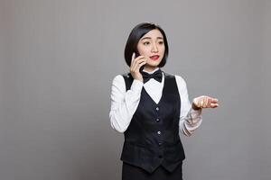 Restaurant woman employee in waitress uniform speaking on smartphone, getting instructions from manager. Attractive asian receptionist chatting on mobile phone, posing studio photo