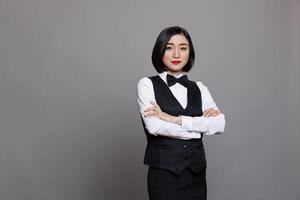 Confident asian restaurant receptionist posing with folded arms and looking at camera with serious face expression. Young waitress with crossed hands wearing black and white uniform portrait photo