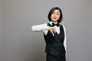 Asian woman receptionist showing thumb down and looking at camera with negative expression grimace on face. Waitress in uniform showcasing service dissatisfaction portrait photo