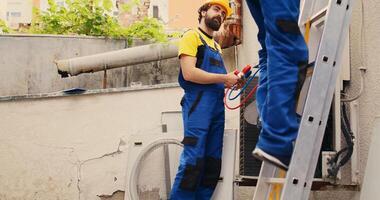 Competent mechanic holding electric hand drill stepping down from folding ladder after finishing servicing rooftop hvac system while colleague assembles manometer to check refrigerant levels photo