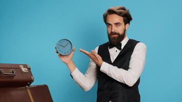 Doorkeeper shows wall clock on camera, checking time and trying to be punctual in hotel travel sector. Man hotel concierge using watch to see hour and minutes in studio next to trolley bags. photo