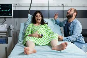 Young couple preparing to be parents in hospital ward bed at healthcare facility. Pregnant woman having painful contractions getting into labor while husband asking for medical assistance photo