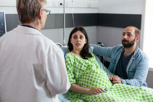 Couple discussing childbirth process with doctor in hospital ward, pregnant woman lying in bed preparing for medical surgery. Patient with pregnancy holding hands on belly being comforting by husband photo