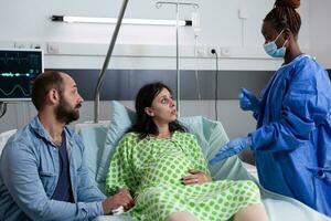 African american surgeron giving medical support to pregnant woman, explaining childbirth surgery in hospital ward. Patient with pregnancy lying in bed having contractions before getting into labor photo