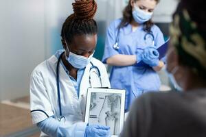 Bone doctor explaining clinical information on tablet to patient during orthopedic medical exam. Orthopedist in modern sterile hospital office wearing face mask and medical gloves photo