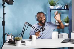 Smiling streamer wearing headphones greeting audience while creating digital content and looking at camera. Cheerful influencer in headset recording video for internet blog photo