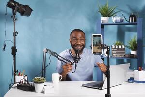 Smiling internet streamer speaking in microphone and looking at smartphone camera while filming video. Cheerful african american vlogger streaming and creating web content using mobile phone photo
