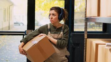 Employee moving stock boxes on racks, listening music on headset while she prepares retail orders for delivery. Young woman working on products shipment, showing dance moves in depot. photo