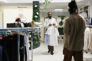 Clothes retail store african american man worker carrying shirts for customer to try. Buyer standing near garment rack and waiting for shop assistant to bring apparel different sizes photo