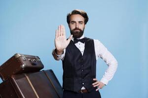 Man raises hand to indicate stop sign against blue background, creating advertisement and expressing rejection gesture on camera. Bellhop doorman presenting his refusal, trolley bags. photo