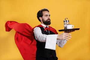 Professional restaurant waiter character holds dish tray and towel while he wears red cape fluttering in studio. Young adult posing with strength and confidence, serving food on platter. photo