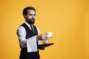 Waiter giving cup of coffee to client on camera, pretending to serve people at fine dining restaurant. Skilled stylish man in formal attire having towel over arm and food platter. photo