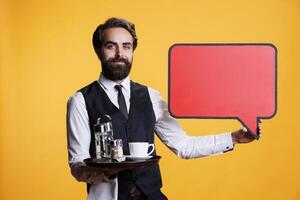 Young employee presents blank speech bubble in studio, working at restaurant and showcasing marketing advertisement with empty billboard copyspace. Waiter shows cardboard icon. photo