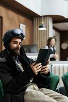 Caucasian man in winter clothing in lounge area, seated comfortably on sofa carrying his phone tablet. Tourist uses device to explore snowboarding slopes, making reservations for winter adventure. photo