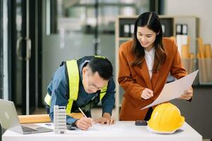Contractor and architect looking at plan, tablet, laptop and talking about working on big project. Building in construction office interior. photo