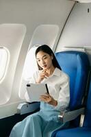 Portrait of  successful Asian businesswoman or entrepreneur in a formal suit on an airplane sitting in business class using a phone, computer laptop. Travel in style, work with grace. photo