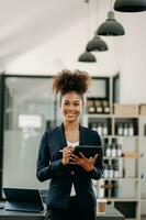 Confident beautiful African businesswoman typing laptop computer and digital tablet while holding coffee at office photo