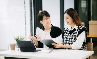 Asian business colleague business presents and explains and using laptop and tablet. Teamwork, financial marketing team, while sitting in modern office room. photo