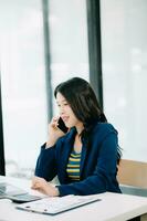 sonriente asiático mujer hablando en el teléfono con un cliente joven positivo hembra contador utilizando teléfono inteligente hablando a equipo a su escritorio relajante con móvil foto